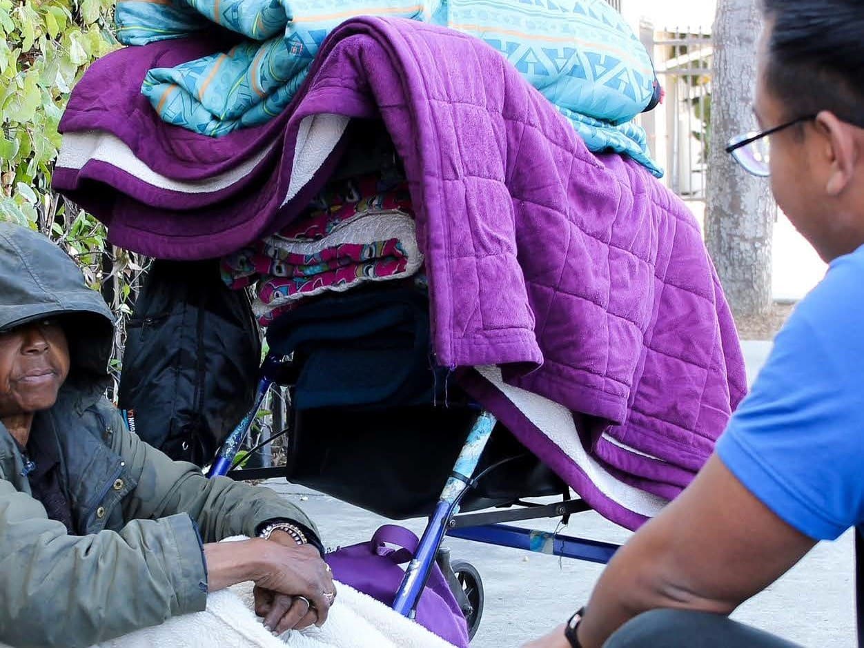 A community outreach moment — two people share a quiet handhold across a stroller in the street, the everyday work of keeping the mission front and center.
